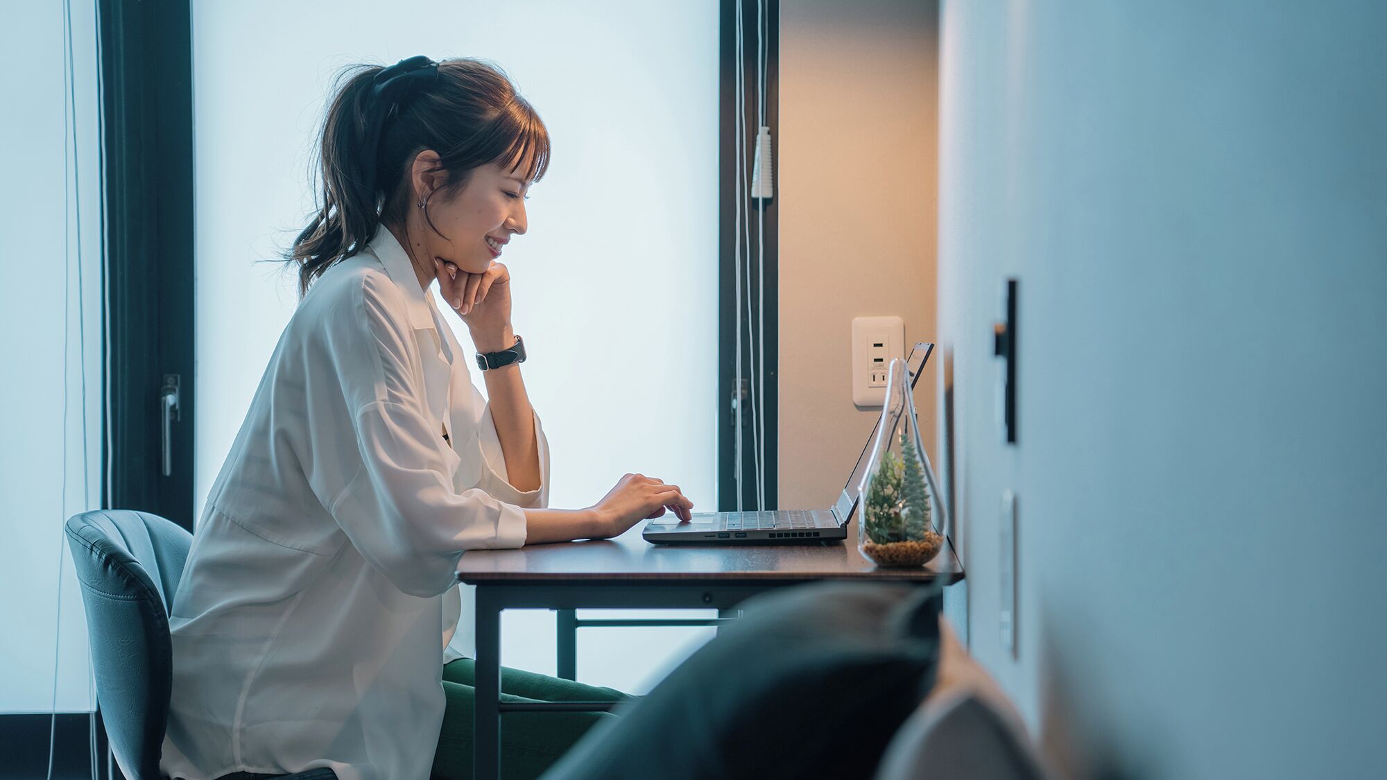 A person working at a desk with a laptop, a power outlet, and a small plant on the table.