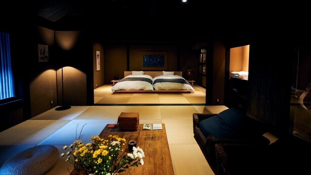 A traditional Japanese-style room with tatami flooring, a low bed with white and gray bedding, a wooden coffee table with flowers, and a dark-toned sofa.