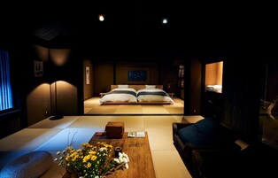 A traditional Japanese-style room with tatami flooring, a low bed with white and gray bedding, a wooden coffee table with flowers, and a dark-toned sofa.