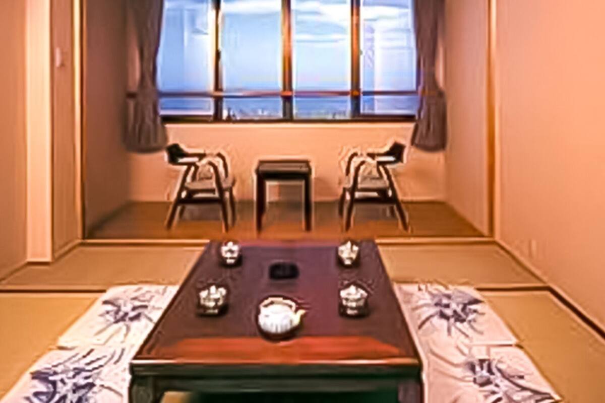 Une pièce de style japonais traditionnel, avec un sol recouvert de tatamis, une petite table sur laquelle est disposé un service à thé, et deux chaises.