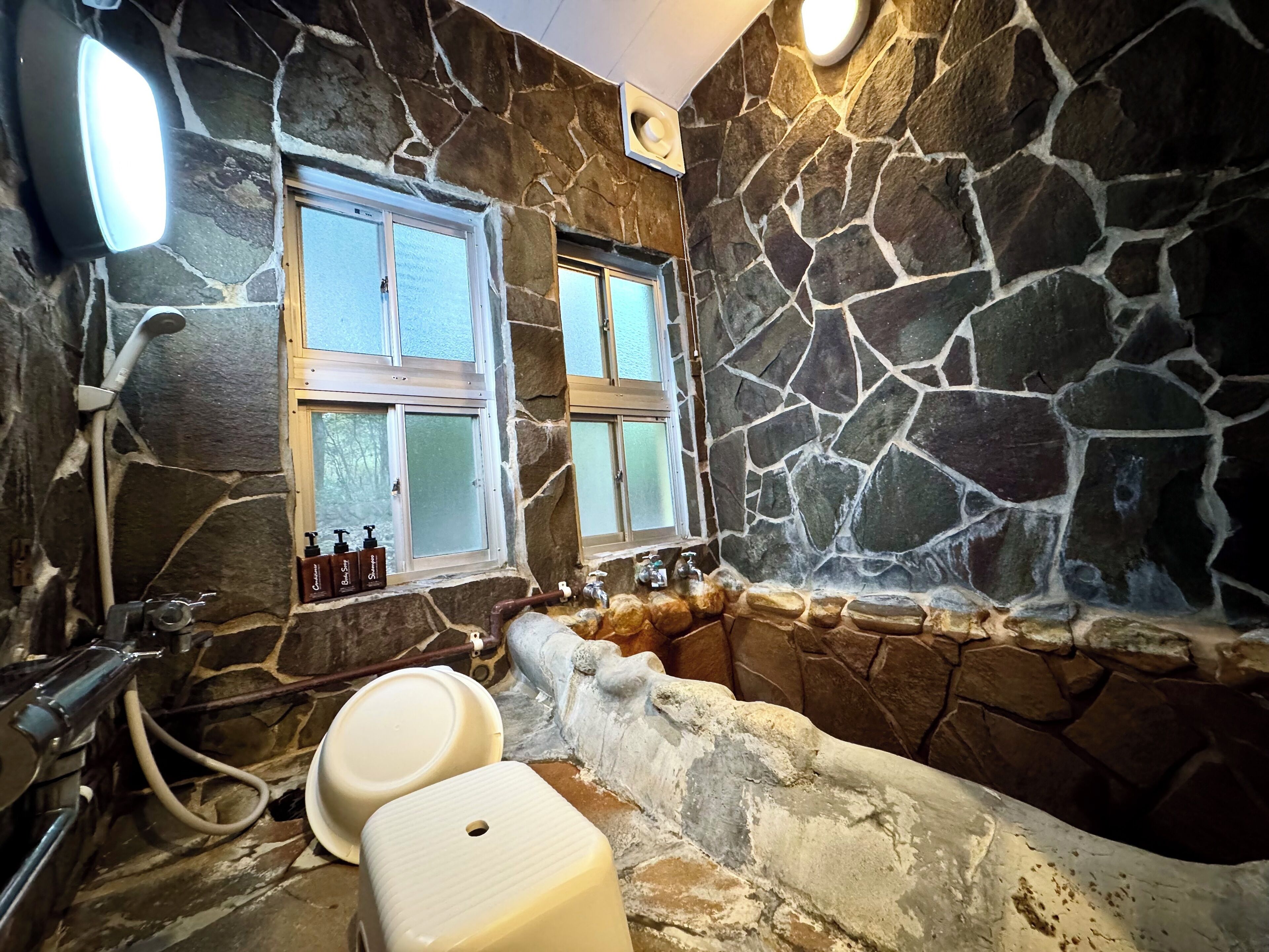 A bathroom with stone walls, a bathtub, a sink, and two windows.