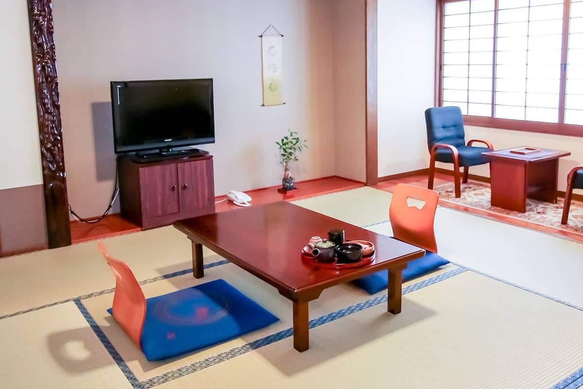 Une pièce de style japonais traditionnel, avec un sol recouvert de tatamis, une petite table avec un service à thé, un téléviseur posé sur un meuble et deux chaises.