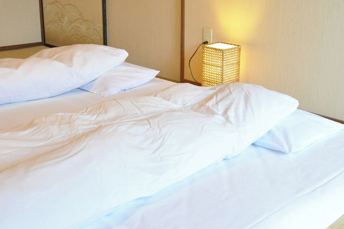 A hotel room with a bed, pillows, and a wall-mounted lamp.