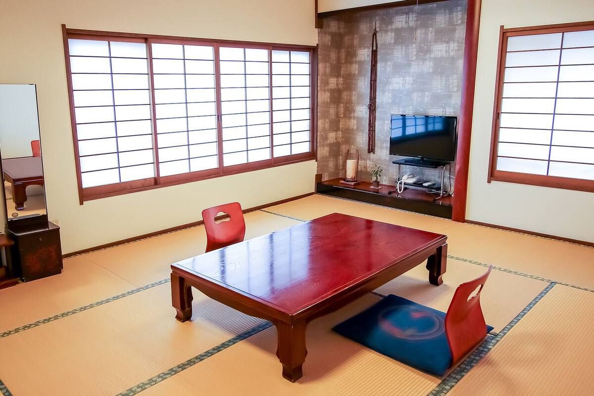 Une pièce de style japonais traditionnel, avec un sol recouvert de tatamis, une table en bois, des chaises rouges, un téléviseur et un miroir.
