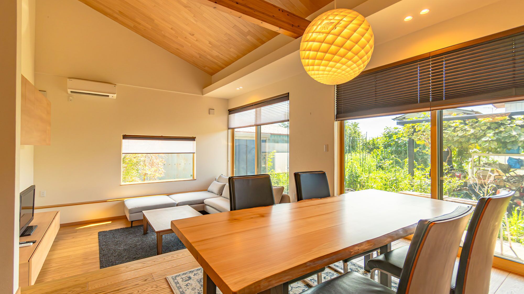 A modern interior with a wooden dining table, chairs, a sofa, and a large window with blinds.