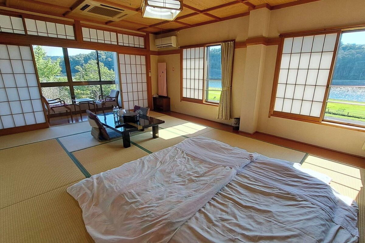 Een traditionele Japanse kamer met tatami vloeren, een futonbed, een lage tafel met stoelen en grote ramen met uitzicht op een meer en bos.