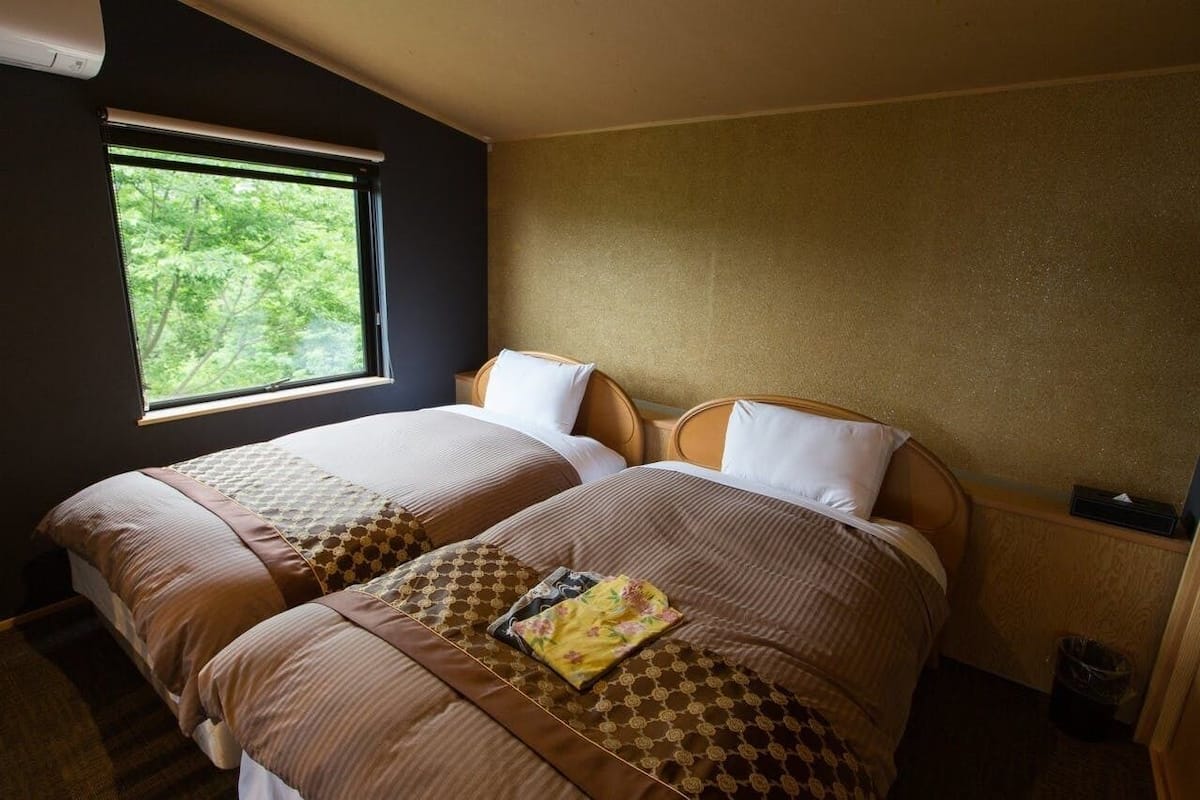 Une chambre avec deux lits, une fenêtre donnant sur des arbres et une petite poubelle.
