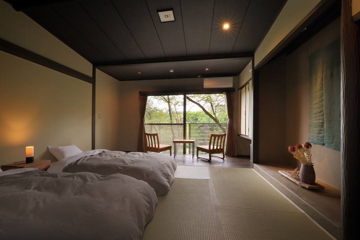 Un dormitorio con dos camas, una mesita, sillas y vistas al exterior.
