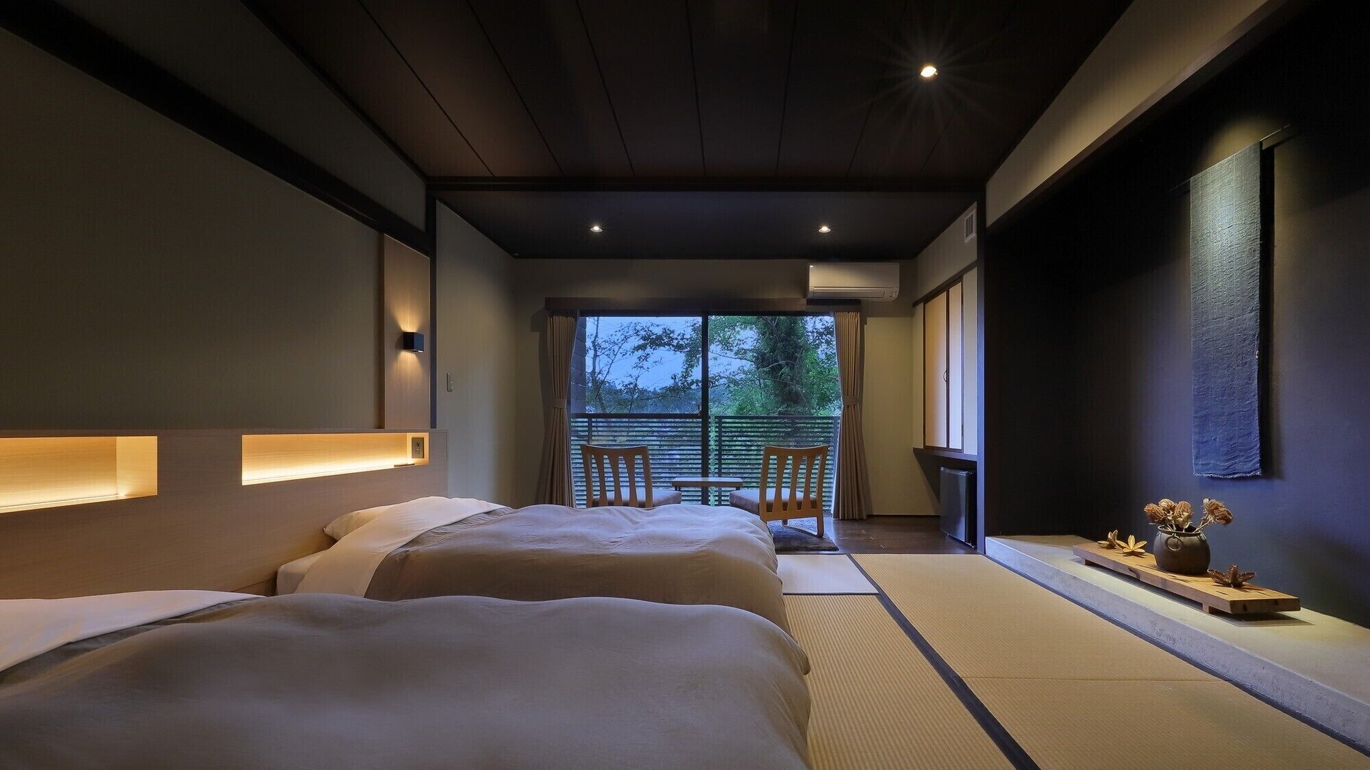 A modern hotel room with two beds, a small table, chairs, a view of the outdoors, and a decorative vase.