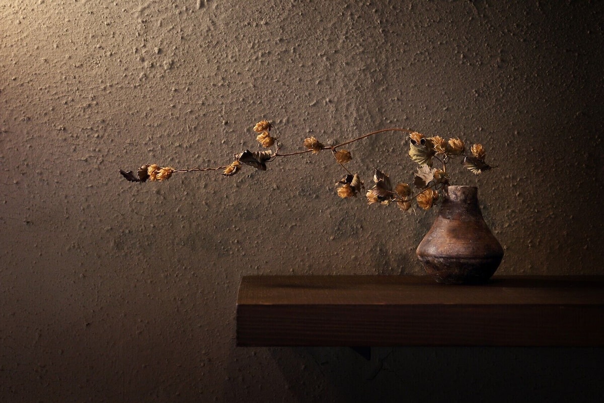 Un jarrón con flores secas sobre un estante de madera frente a una pared con textura.