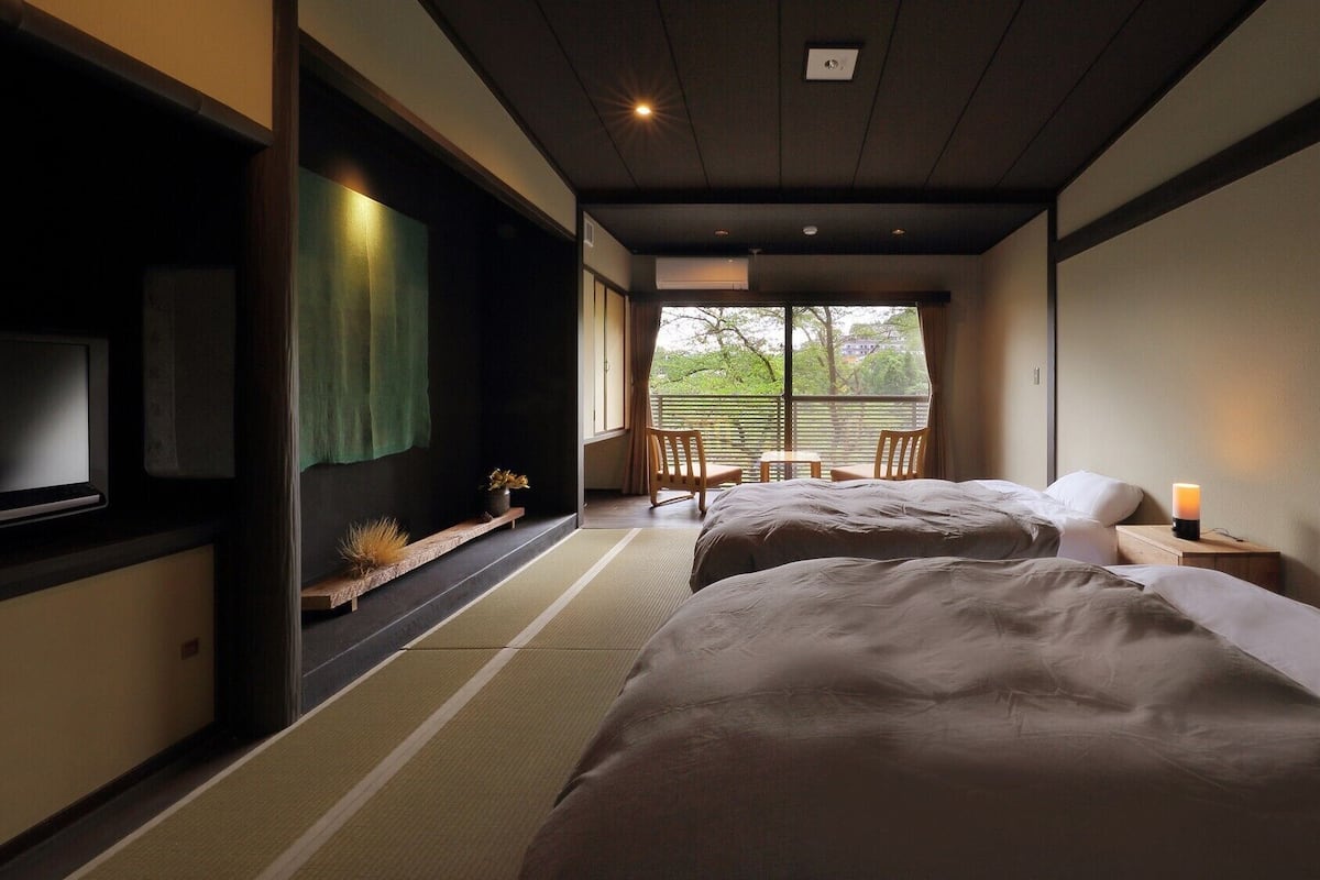 Habitación de hotel con dos camas, televisión, ventana con vistas y una pequeña mesa con sillas.