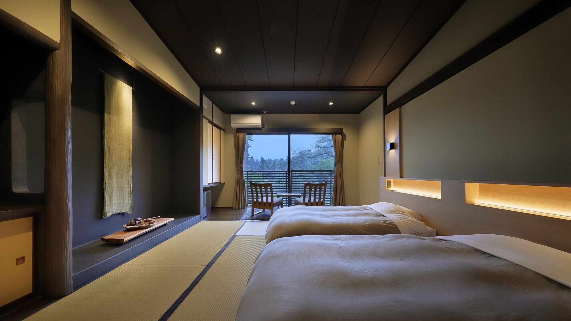 A hotel room with a bed, a small table, chairs, a view of trees, and a sliding door.