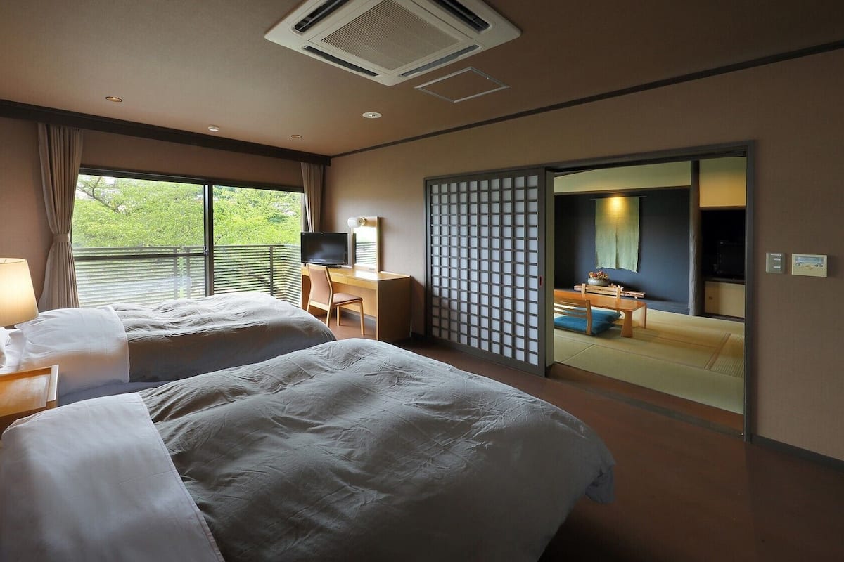 Habitación de hotel con dos camas, un escritorio con una silla, un televisor, una puerta corredera y vista a zonas verdes.