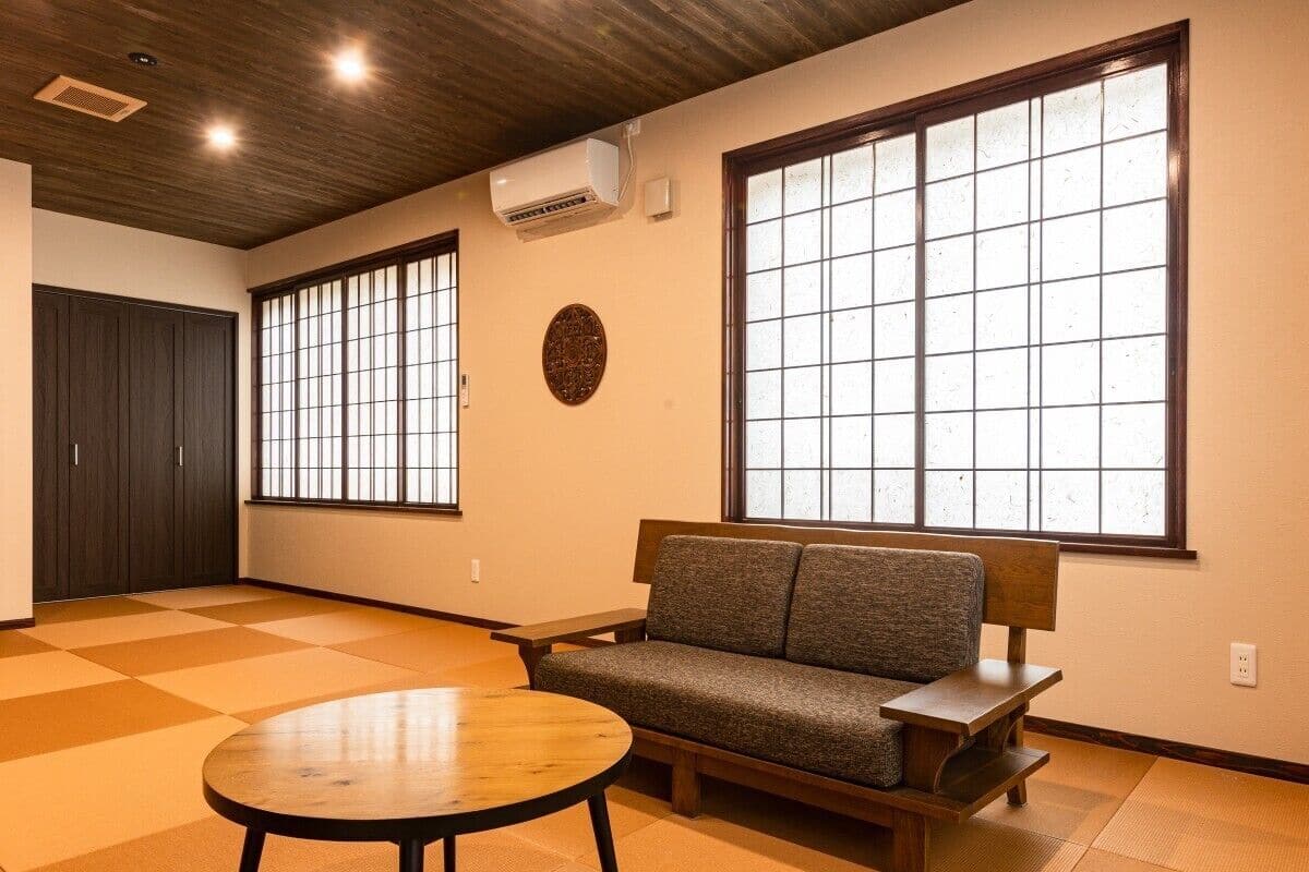 Ein Zimmer mit Tatami-Böden, einer Couch, einem kleinen Tisch und großen Fenstern mit Gittermustern.