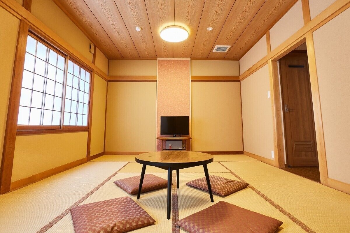 Ein traditioneller japanischer Raum mit Tatami-Böden, einem Tisch aus Tatami-Matten, einem Fernseher auf einem niedrigen Schrank und einer Schiebetür.