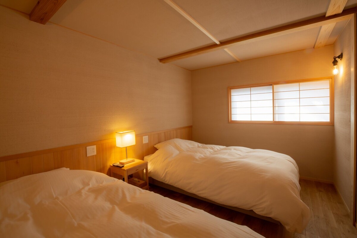 Quarto com duas camas, mesa de cabeceira com abajur, janela com persianas e vigas de madeira no teto.