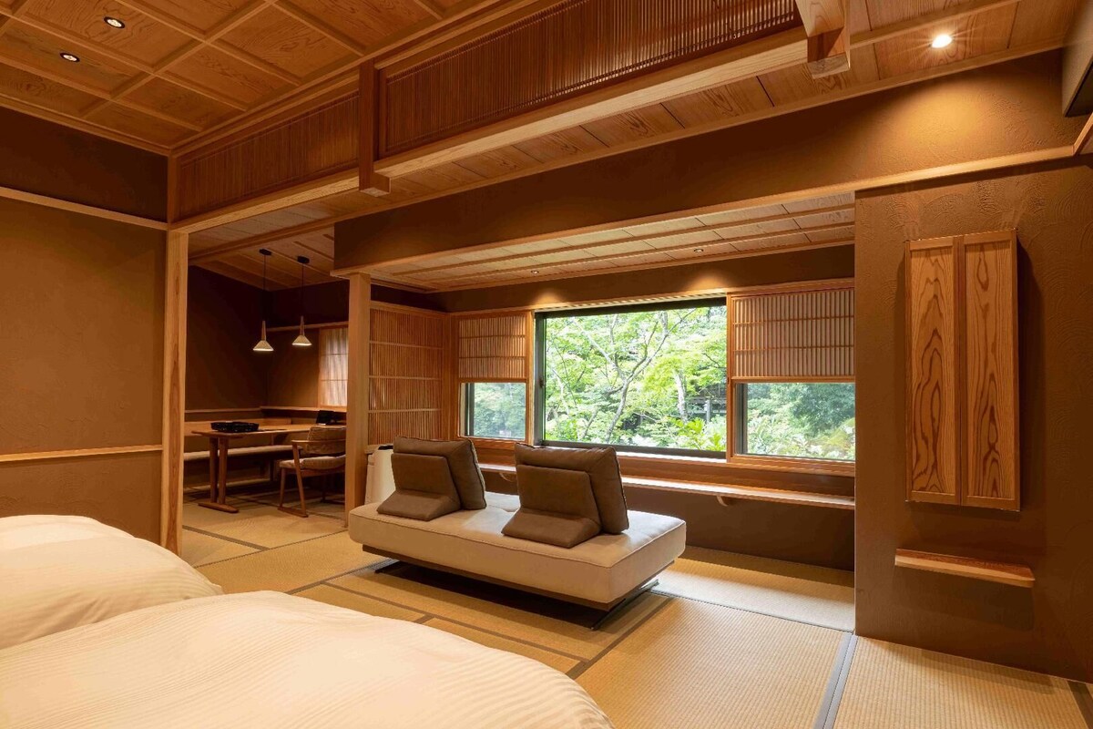 Quarto com sofá, mesa de jantar e vista para o verde pela janela.