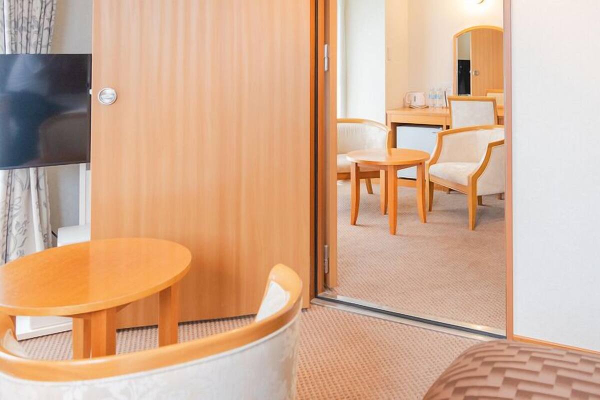 A hotel room with a wooden door, a small round table, two chairs, a bed, and a mirror on the wall.