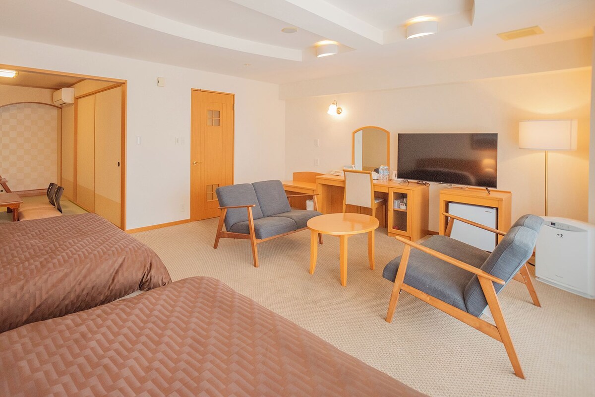 A hotel room with a bed, two armchairs, a small table, a desk with a TV, and a mirror.