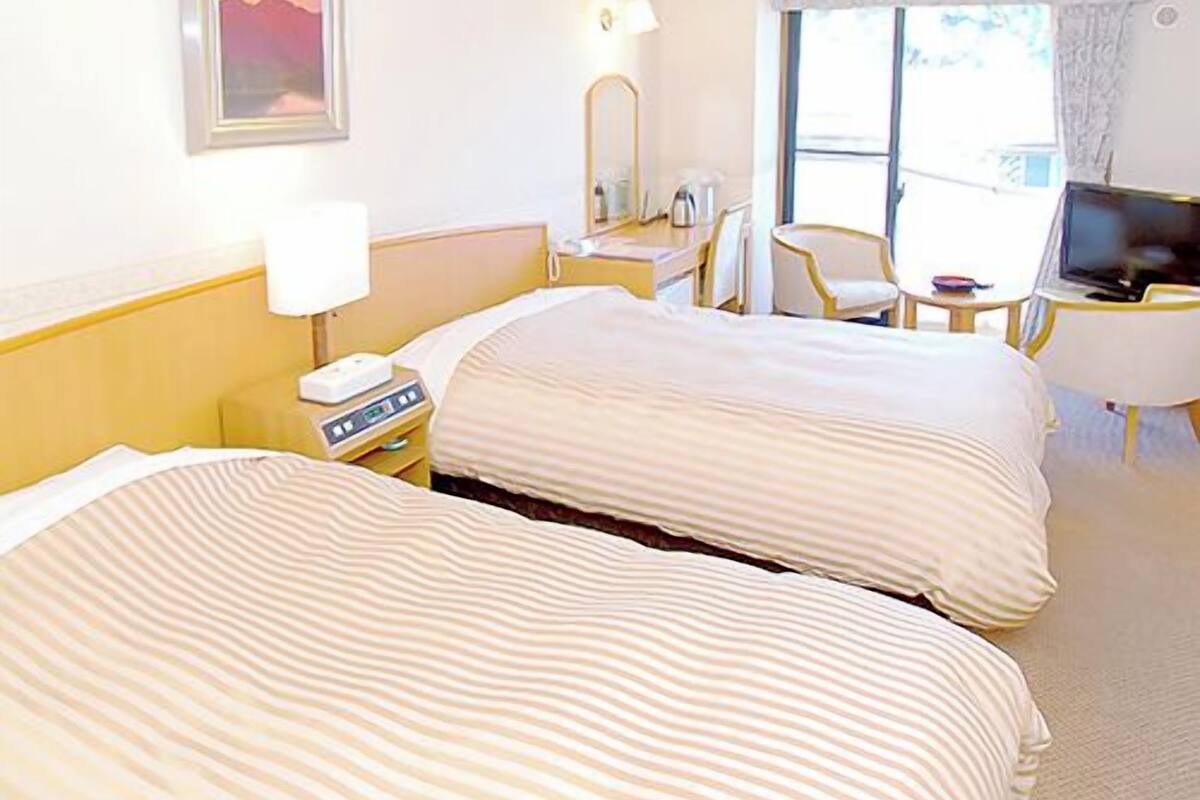 A hotel room with two beds, a TV, a desk with a lamp, a chair, and a window with curtains.
