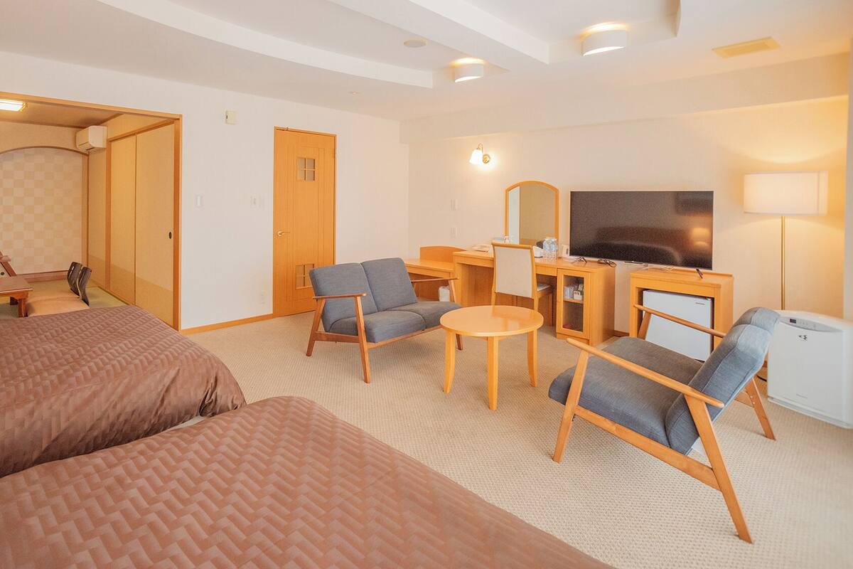 A hotel room with a bed, two armchairs, a small table, a desk with a TV, and a mirror.