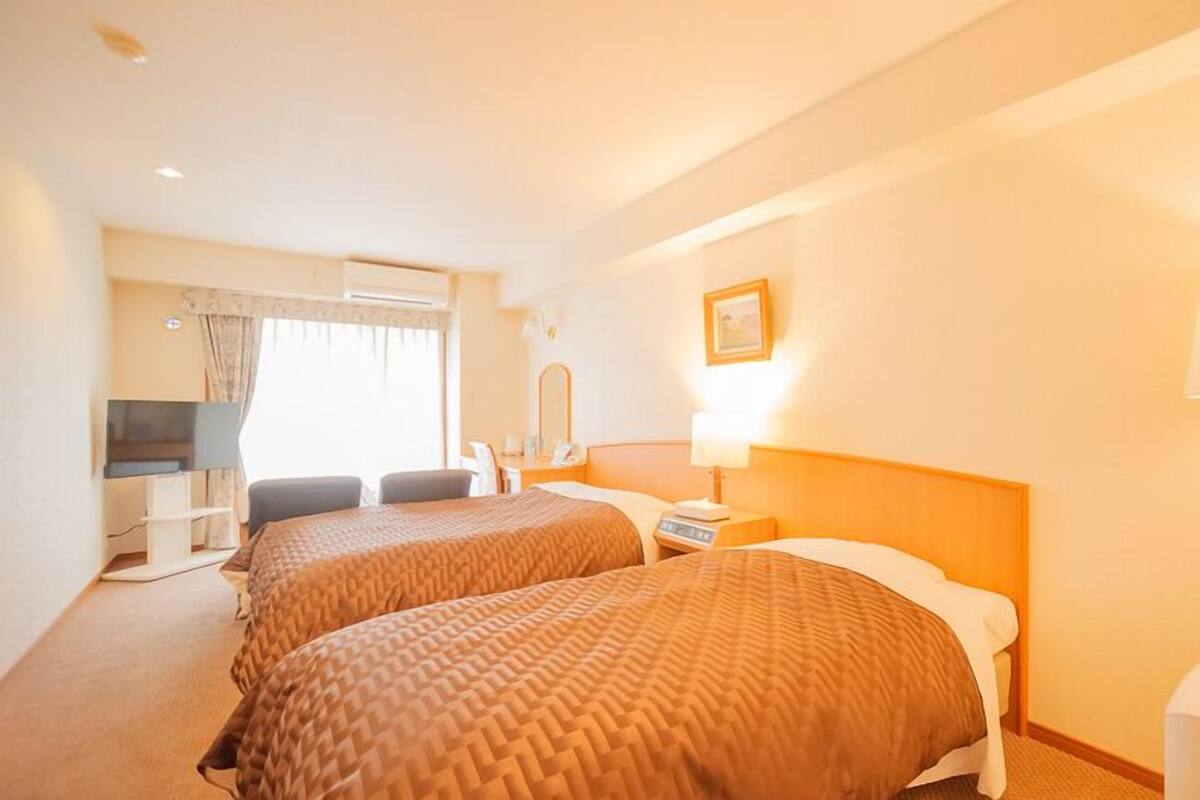 A hotel room with two beds, a desk, a chair, a TV, and a lamp.