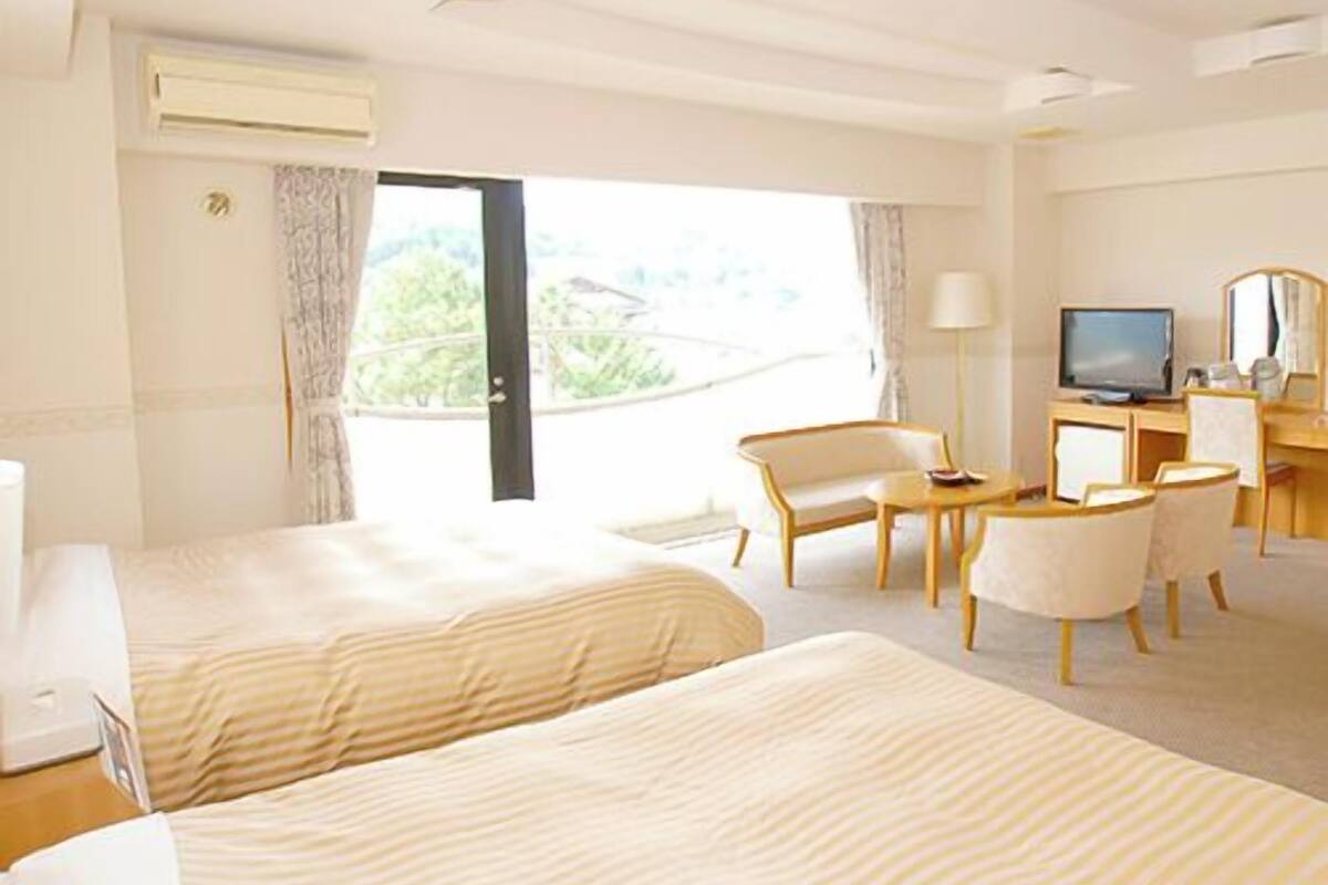 A hotel room with two beds, a desk with a television, a chair, and a view of greenery outside.