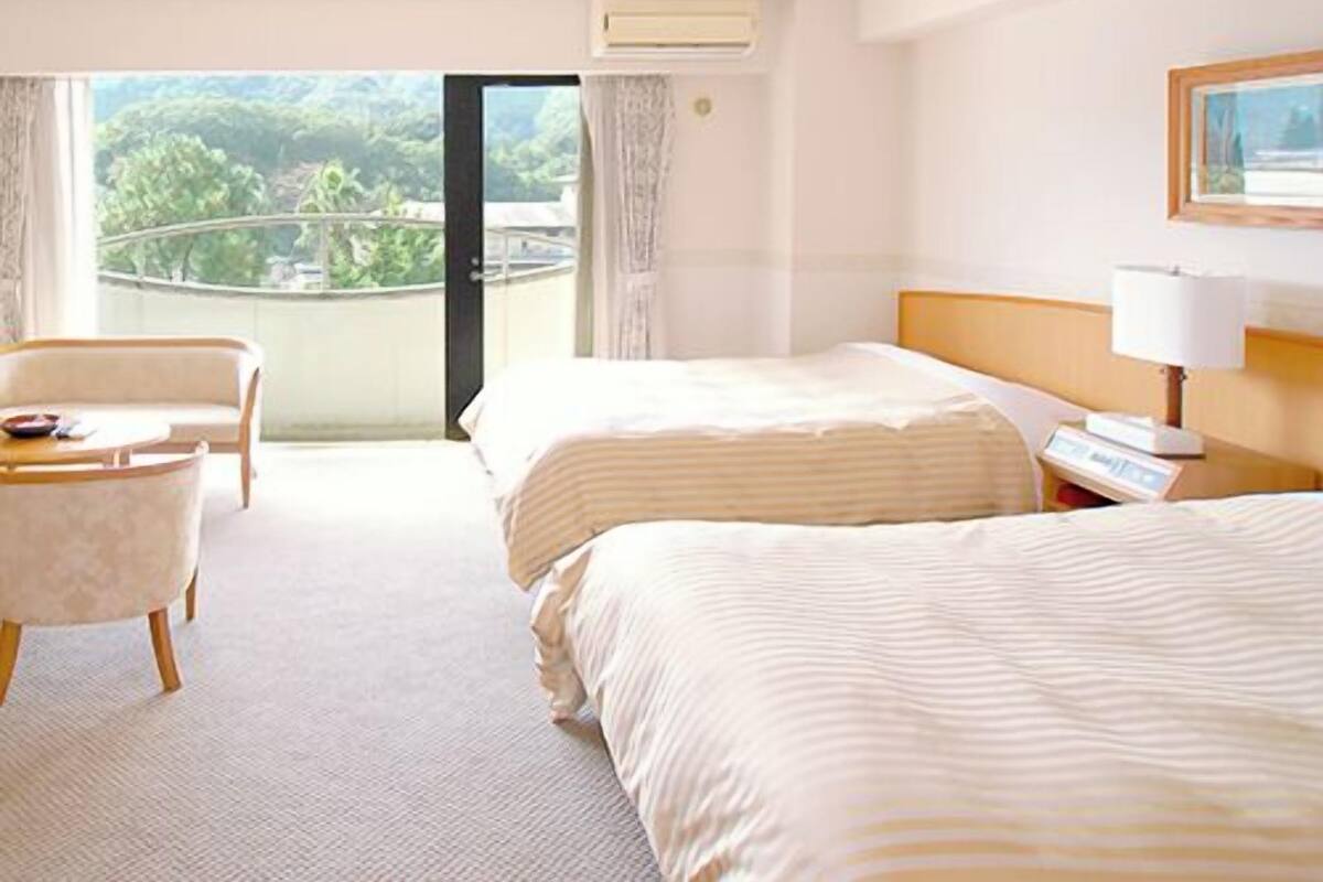 A hotel room with two beds, a sofa, a desk, and a view of greenery outside.