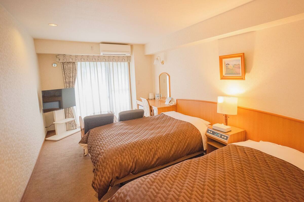 A hotel room with two beds, a desk with a chair, a TV, and a window with curtains.