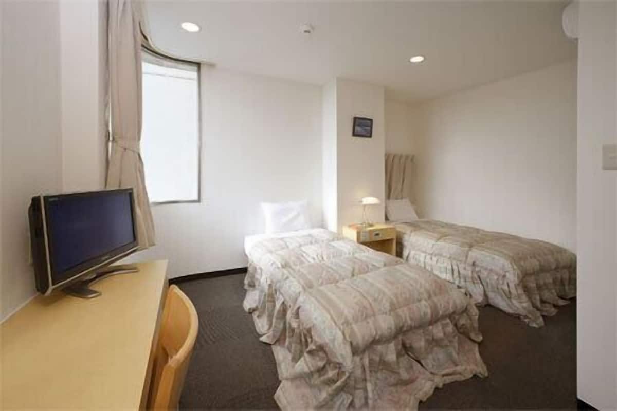Ein Hotelzimmer mit zwei Betten, einem Schreibtisch mit Computer, einem Fenster mit Vorhängen und einem kleinen Nachttisch mit Lampe.