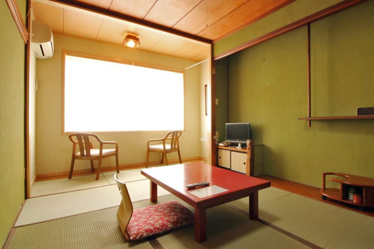 Une pièce de style japonais traditionnel, avec un sol recouvert de tatamis, une basse table en bois, deux chaises, un téléviseur et des portes coulissantes.