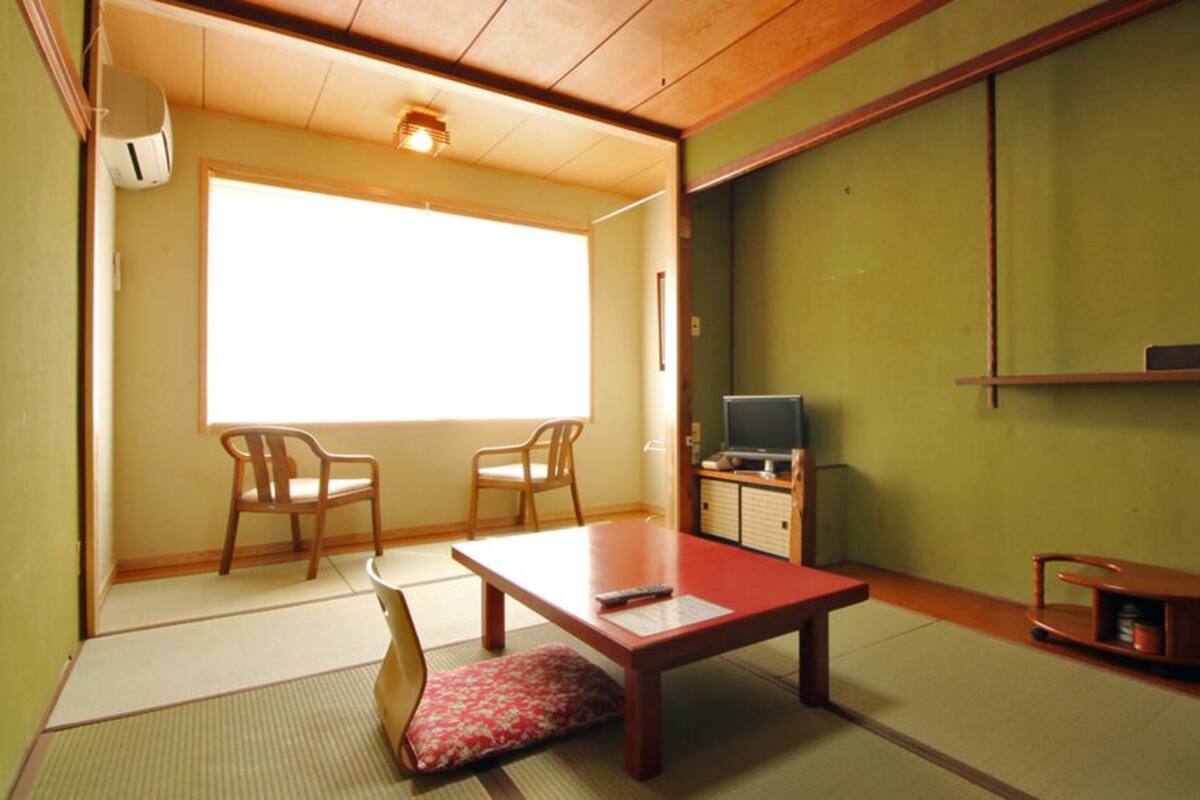 Una habitación japonesa tradicional con suelo de tatami, una mesa de madera baja, dos sillas, un televisor y puertas correderas.