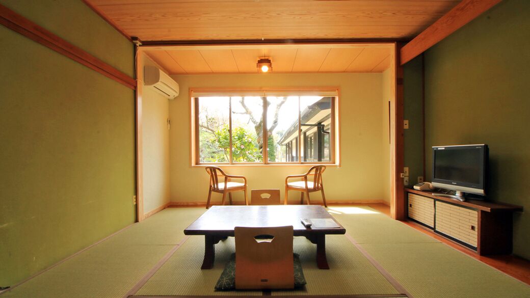 Ein traditioneller japanischer Raum mit Tatami-Böden, einem niedrigen Tisch, zwei Stühlen, einem Fernseher und einem Fenster mit Blick auf einen Baum und ein Gebäude.