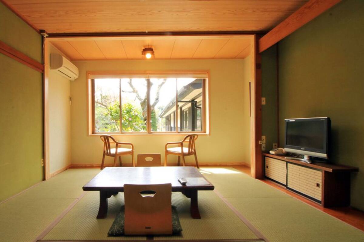 Une pièce de style japonais traditionnel, avec un sol recouvert de tatamis, une table basse, deux chaises, un téléviseur et une fenêtre donnant sur un arbre et un bâtiment.