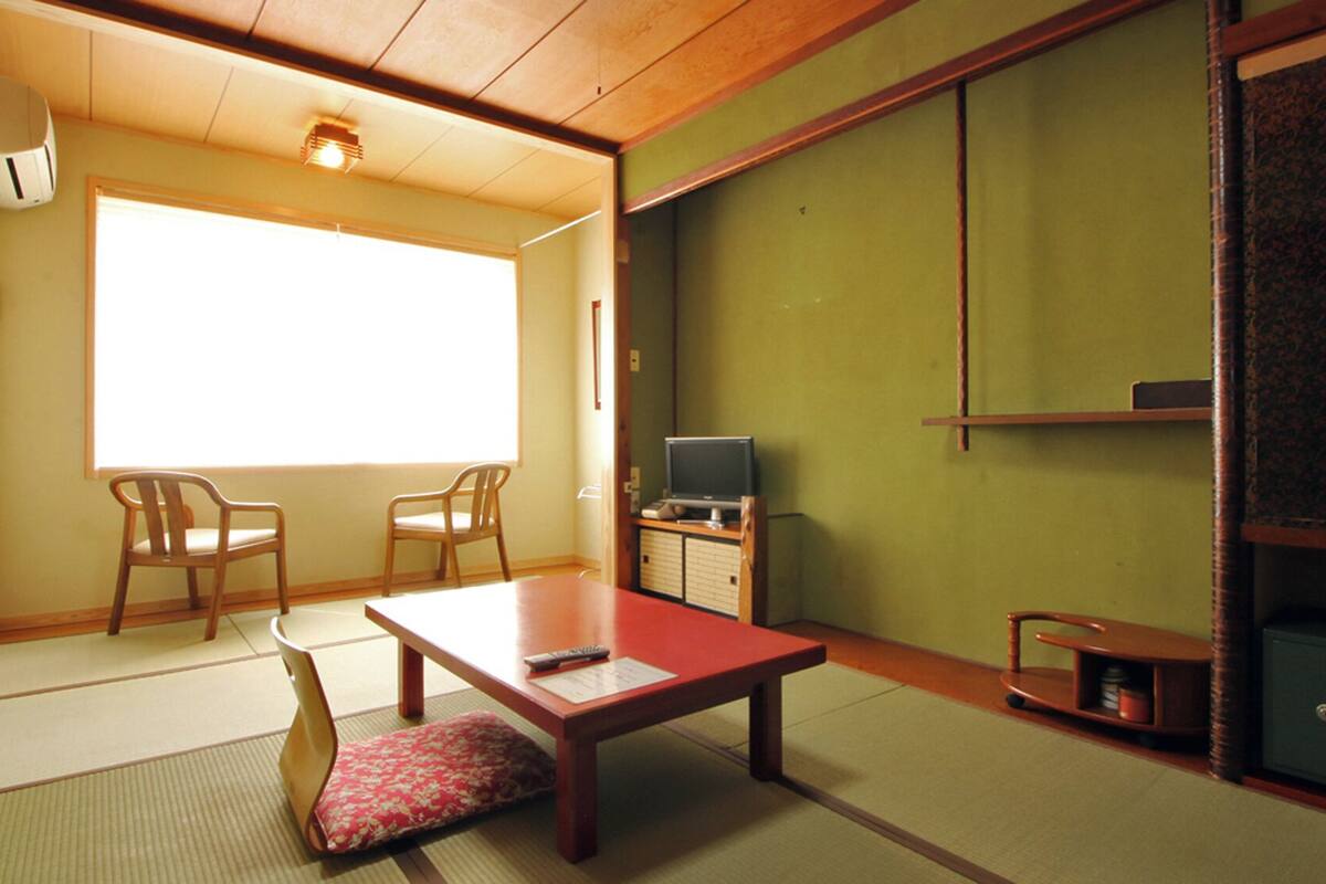 Une pièce de style japonais traditionnel, avec un sol recouvert de tatamis, une table basse, deux chaises, un téléviseur et une porte coulissante.