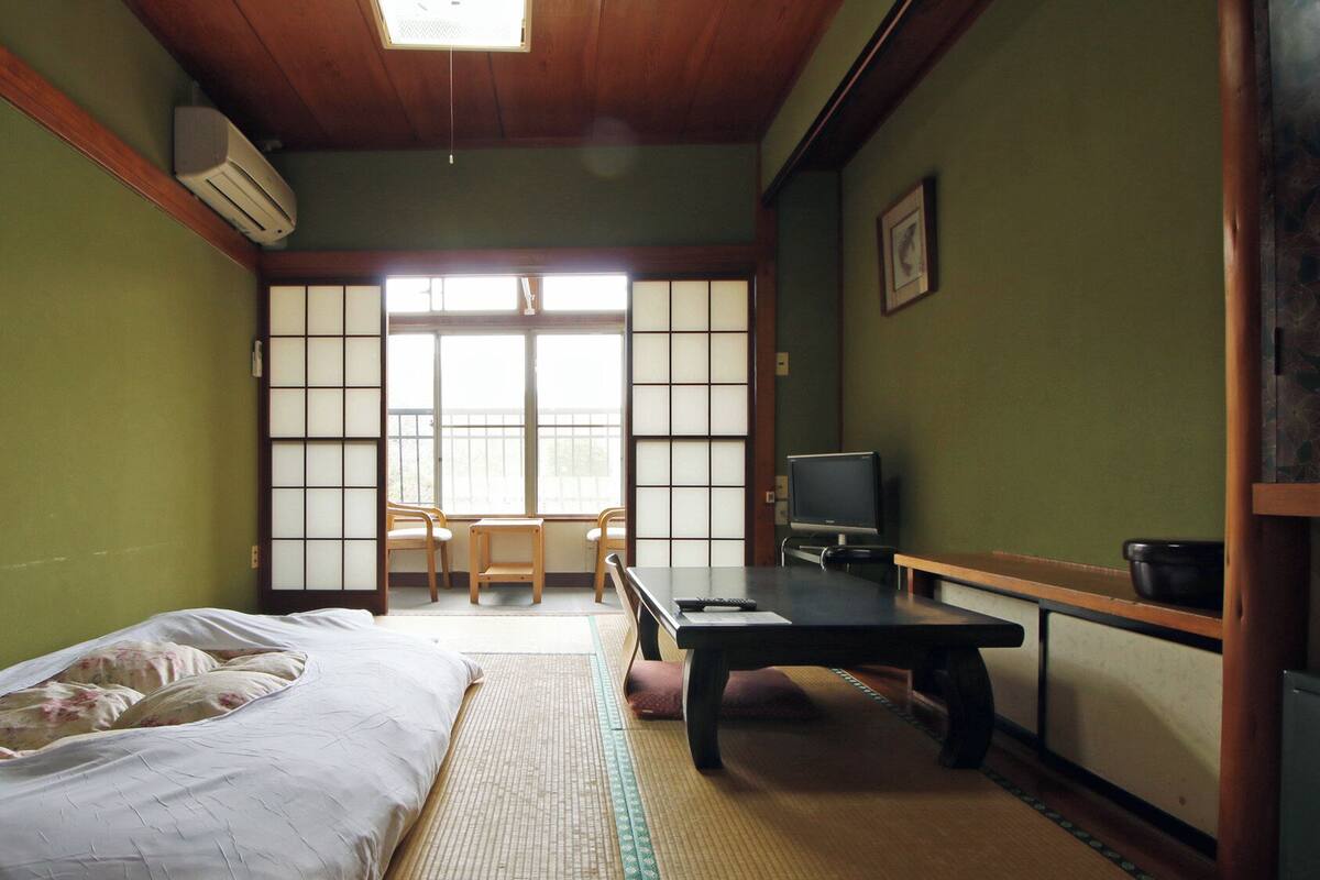 Une pièce de style japonais traditionnel, avec un sol recouvert de tatamis, un futon servant de lit, une télévision et une porte coulissante donnant sur un balcon.