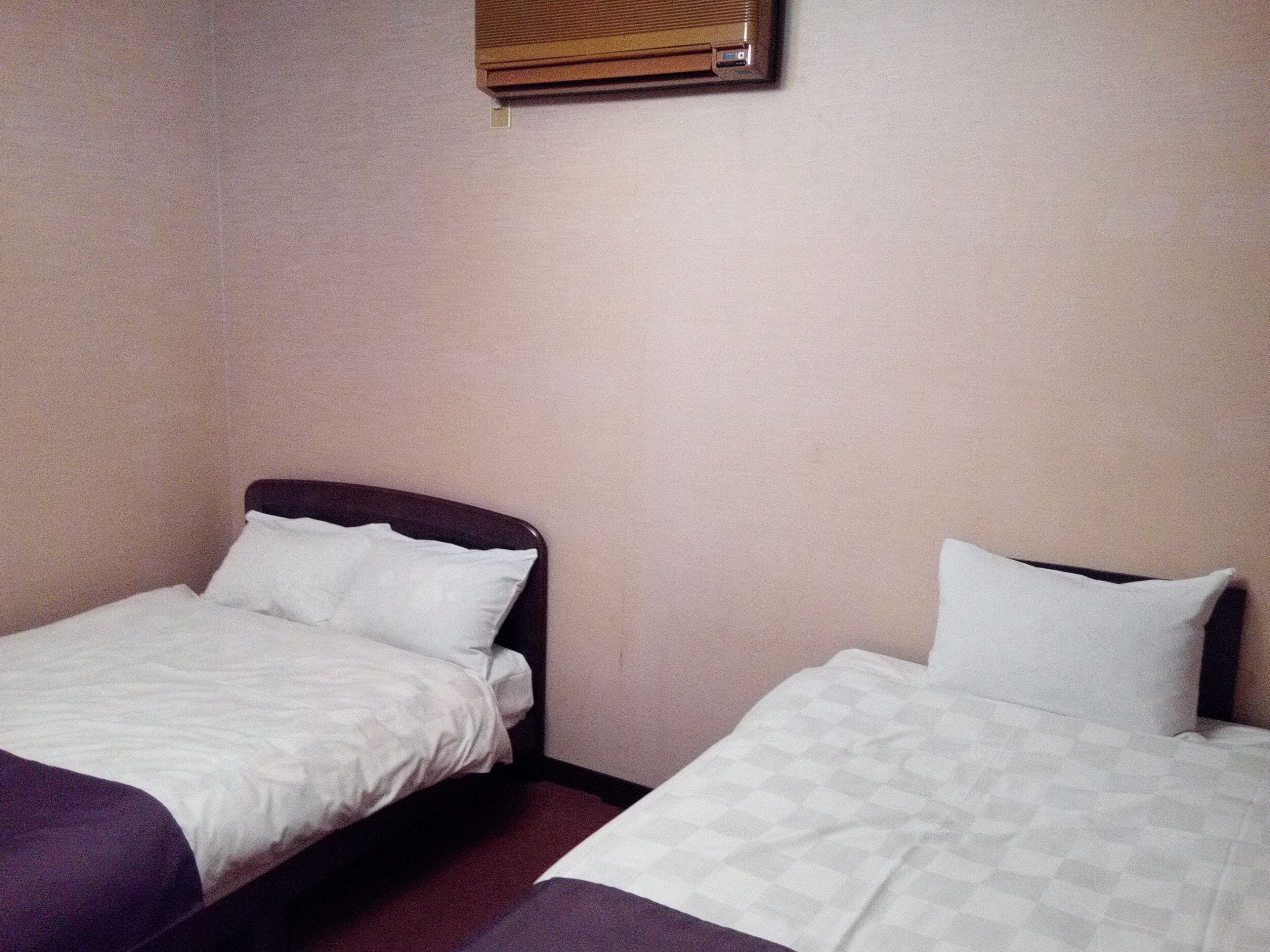 Dos camas individuales con sábanas blancas en una habitación con paredes color beige y una ventana con persianas.