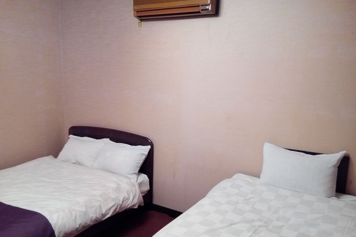 Dos camas individuales con sábanas blancas en una habitación con paredes color beige y una ventana con persianas.