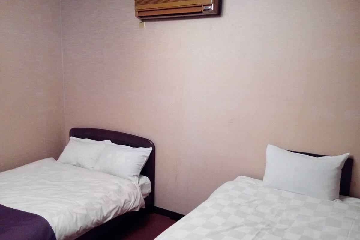 Deux lits simples avec du linge de lit blanc, dans une chambre aux murs de couleur beige et dotée d’une fenêtre avec des stores.