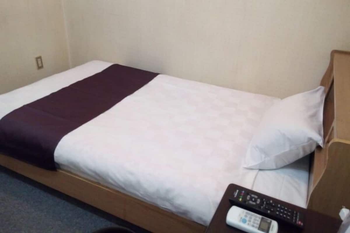 Una cama individual con una colcha blanca y morada, un cabecero de madera y un control remoto sobre una mesita de noche de madera.