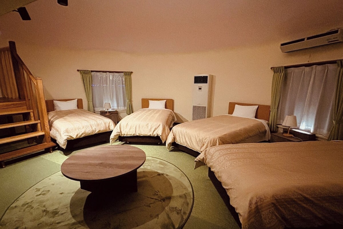 Ein Hotelzimmer mit vier Betten, einer hölzernen Bank, einem runden Tisch und einer Klimaanlage.
