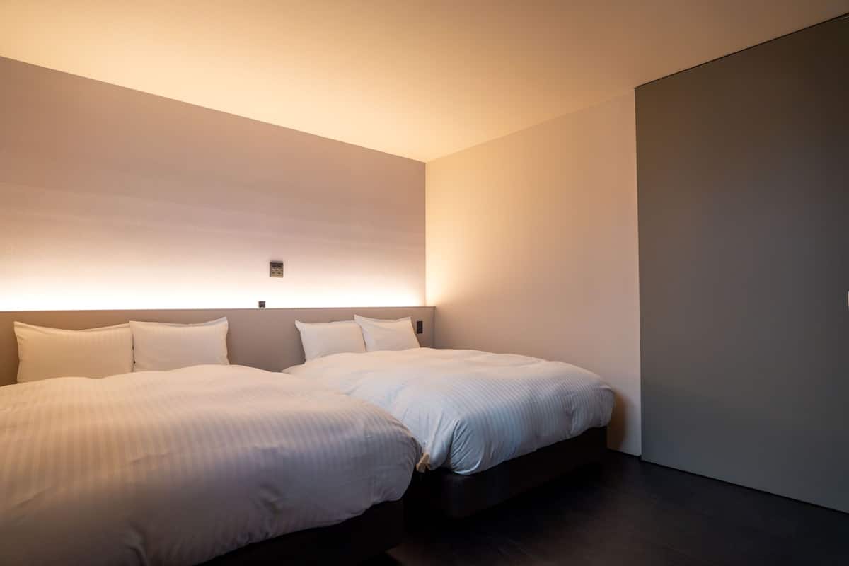 Une chambre d’hôtel moderne avec deux lits, un plancher foncé et un luminaire fixé au mur.