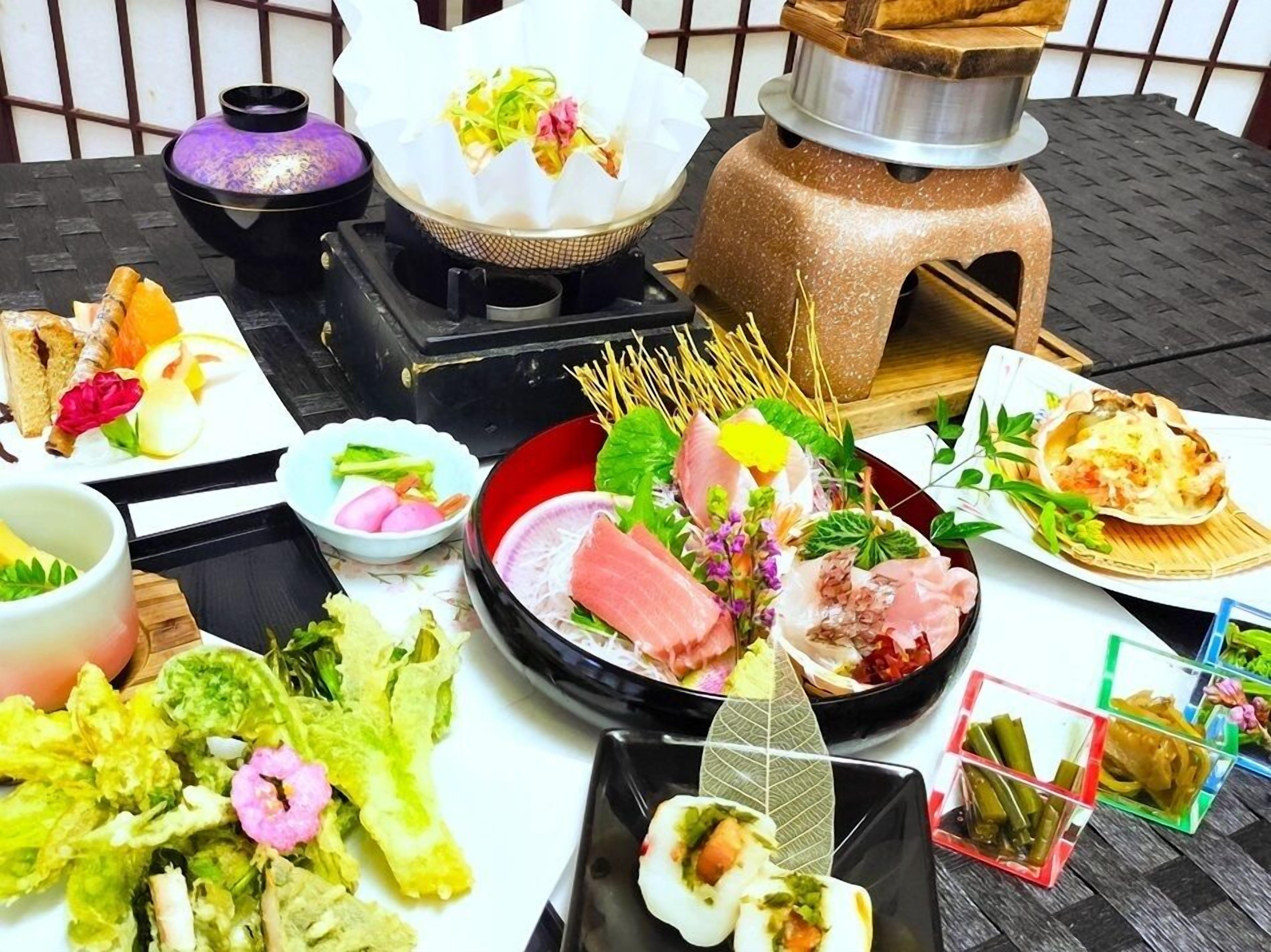 Forskellige japanske retter, blandt andet sashimi, tempura og sushi, anrettet på et bord med et traditionelt japansk tesæt og en kulgrill.
