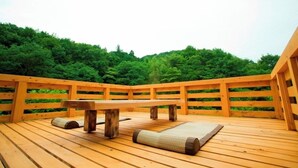 A wooden deck with a bench and a mat, surrounded by a wooden railing, overlooking a forested area.