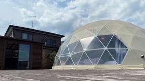 A geodesic dome structure with a transparent roof, adjacent to a modern building with a wooden facade and a visible antenna.