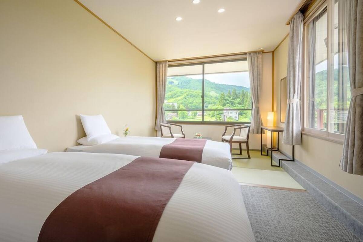 A hotel room with two beds, a small table, chairs, and a large window with a view of greenery.