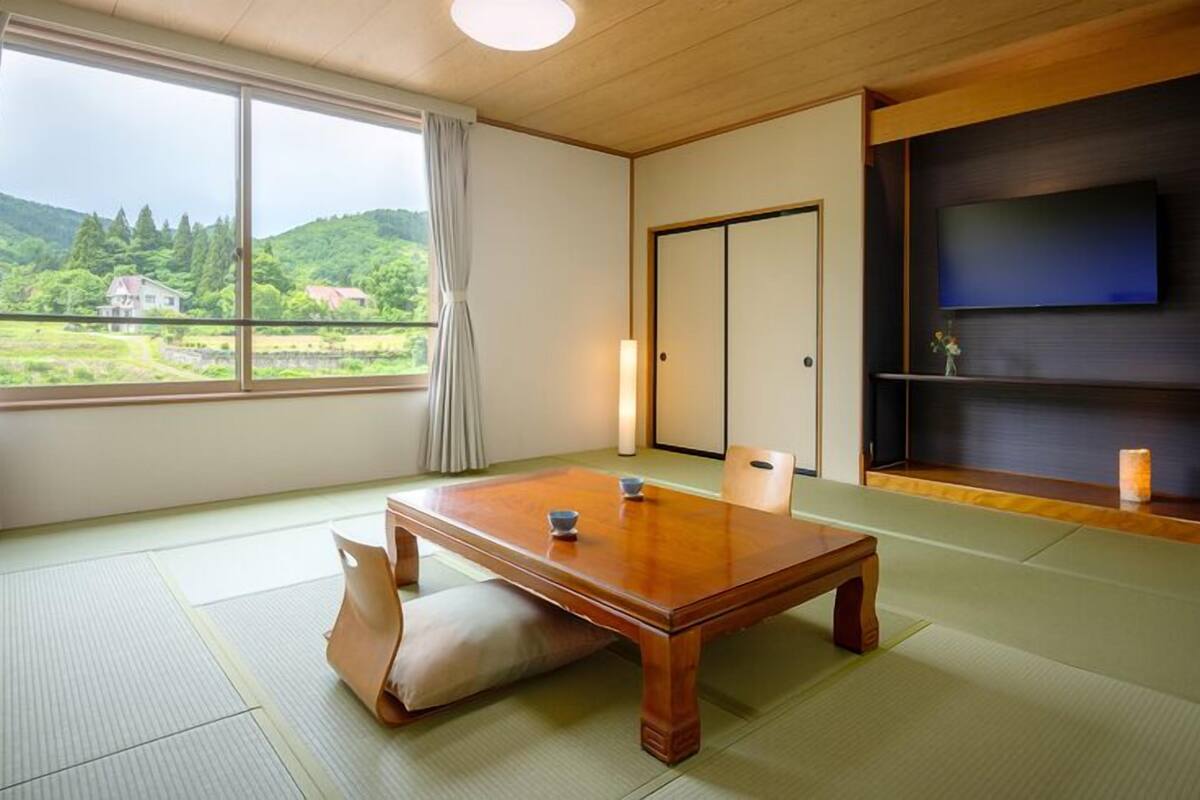 A traditional Japanese room with tatami flooring, a low wooden table, a sliding door, and a TV on a dark wall.