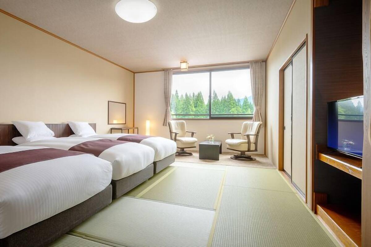 A hotel room with two beds, a TV, a window with a view of trees, and a tatami mat floor.
