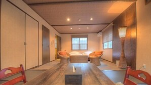 Una habitación con una larga mesa de madera, dos zonas de estar con cojines, una cama y una lámpara alta.