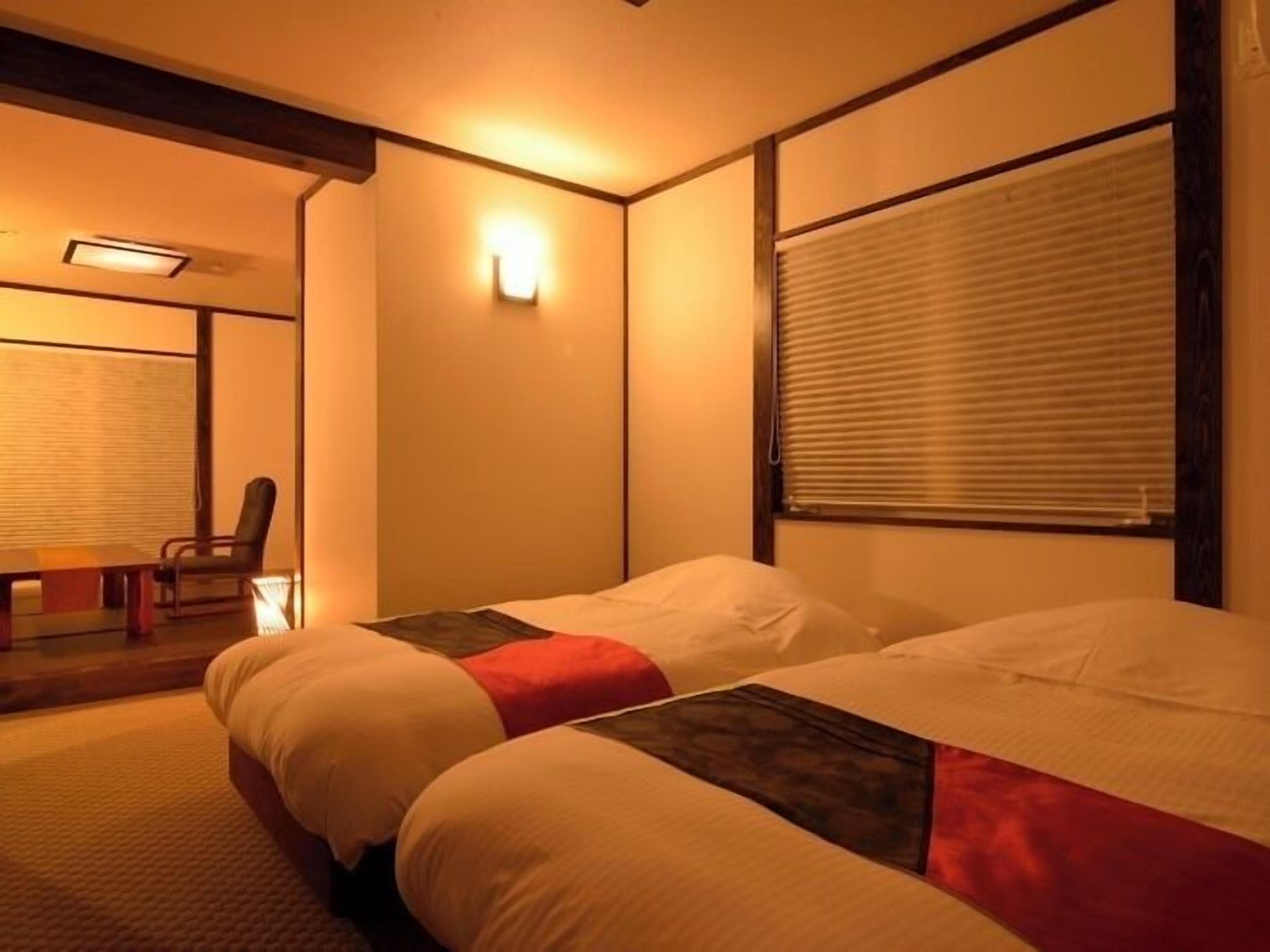 A hotel room with a bed, a chair, a table, and a window with blinds.